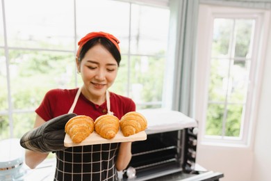 young smiling asian female baking croissant bread in oven, bakery cooking concept with copy space