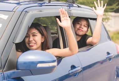 two teenage girls do a friendly wave while passing by and driving in a car. waving hello and going on their 1st trip. a novice driver with a student license permit.