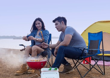 young asian couple grilling barbecue in front of tent while camping at lakeside