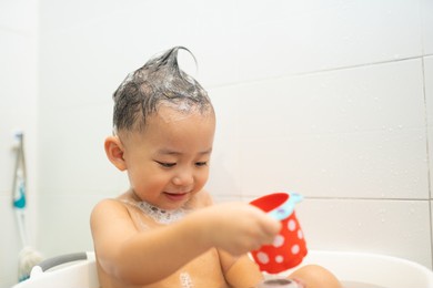 smiling asian baby child boy play with toy at bathtub in bathroom, cute asian toddler boy with funny hair, a child bathes, lying in a white bath with foam from soap and shampoo.shower soap skin care.