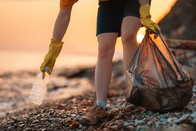 earth day. cleanup garbage on the ocean coast. a volunteer with polyethielene bag picking up a plastic bottle on the beach. close-up. sunset in the background. the concept of conservation of ecology.