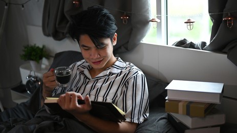 happy asian man drinking coffee and reading book on his bed in the morning.