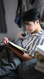 young asian man drinking coffee and reading book on bed in the morning.