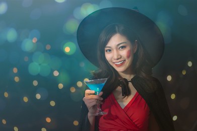 portrait of beautiful young asian woman in witch halloween costumes drinking cocktail in halloween party over dark magic background - holiday party concept
