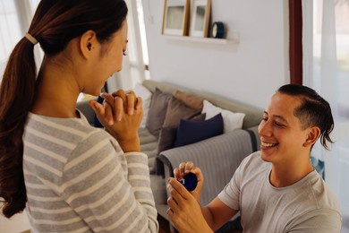 young man proposing his girlfriend with engagement ring at home. asian woman getting surprised by her boyfriend with marriage proposal