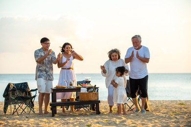 happy asian family on holiday vacation. group of multi generation family little girl with parents and grandparents relax and enjoy with dinner party and dancing together on the beach at summer sunset.