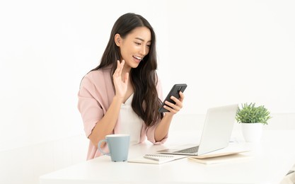 portrait of young business asian woman uses mobile phone for vdo call via 5g internet connection smile wave hand hello. during break from working in modern office. mobile business technology concept