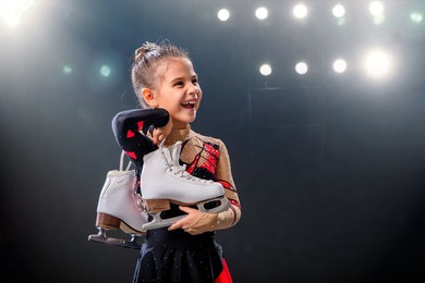 portrait of a happy little skater with skates in her hands on the ice arena, dark background