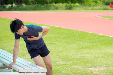 an asian young man feeling chest injury and pain (heart attack) near the racetrack at the stadium after exercise, with copy space for text or design. concepts of health, sports healthcare and medical.