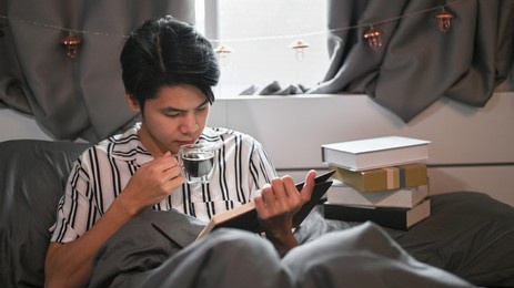 man drinking coffee and reading a book on his bed in the morning.