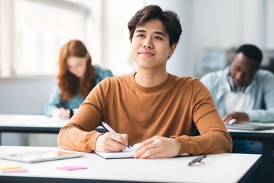 school's in session. portrait of cheerful happy asian male student sitting at table in academic auditorium, listening to tracher. higher educational institutions, learning with pleasure concept