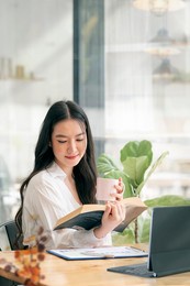 portrait of young beautiful woman holdling mug and reading a book while relaxing during work at home. vertical view.