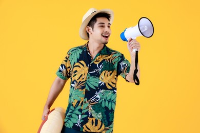 handsome young asian man in hawaiian shirt speaking into megaphone. half body studio portrait on yellow background. relaxed and happy people in summer holidays concept