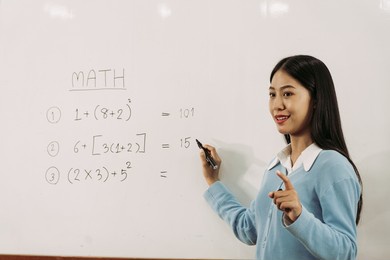 asian female teacher is teaching students at the classroom while pointing at numbers on the white board.