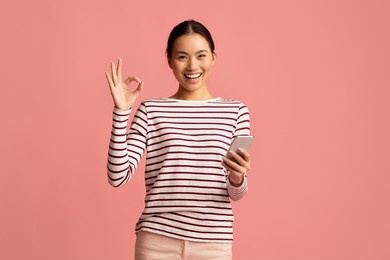 mobile banking. happy young asian woman holding smartphone and showing ok gesture, cheerful korean female enjoying making payments online while standing isolated over pink background, copy space