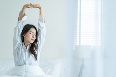 asian beautiful girl in pajamas wake up in the morning with happiness. attractive young woman smiling, feel happy and relax then stretching body after getting up from sleep on bed in bedroom at home.