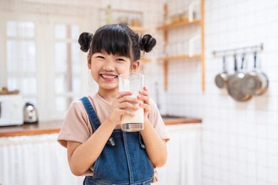 portrait of asian little cute kid holding a cup of milk in kitchen in house. young preschool child girl or daughter stay home with smiling face, feel happy enjoy drinking milk and then look at camera.