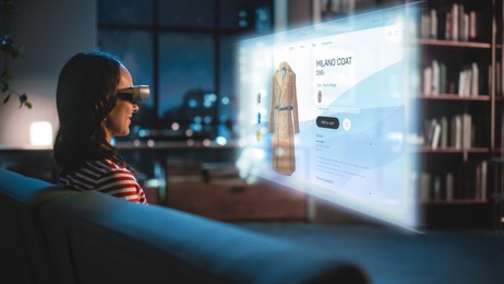 young woman using virtual reality headset at home, sitting on a couch, shopping online via vr clothing store. evening resting at apartment, choosing new look. over the shoulder