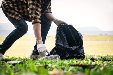 volunteer man in gloves walking and stooping to collect plastic bottles into plastic black bag for cleaning the park during environmental activity to collecting garbage