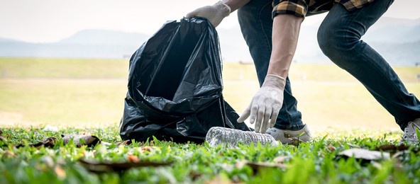 volunteer man in gloves walking and stooping to collect plastic bottles into plastic black bag for cleaning the park during environmental activity to collecting garbage