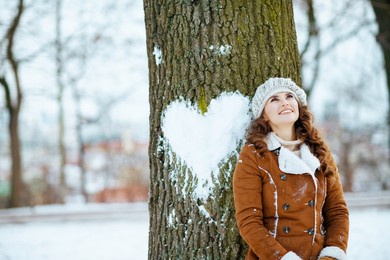 happy modern 40 years old woman in a knitted hat and sheepskin coat near tree with snowy heart outdoors in the city park in winter.