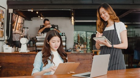 cheerful asian lady waitress with notebook taking receive order menu from young client girl at urban cafe. young asian freelance women working on laptop at coffee shop. owner small business concept.
