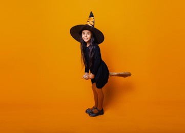 little girl dressed as a halloween witch in a black dress and hat flies on a broomstick on a yellow background.