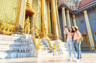 happy asian women friends traveler with camera sightseeing in temple of the emerald buddha, wat phra kaew, popular tourist place in bangkok, thailand