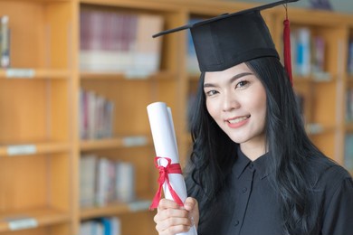 portrait of a young asian university female graduate in graduation gown and mortarboard celebrates diploma degree looking at camera with book self in background