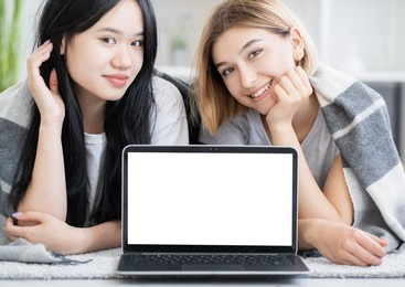 online movie. happy female friends. pajamas party. digital technology. pretty smiling asian caucasian women laying floor with laptop blank screen light room interior.