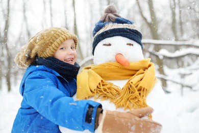 little boy building snowman in snowy park. child embracing snowman wearing hat and scarf. active outdoors leisure with family with children in winter. kid during stroll in a snowy winter park