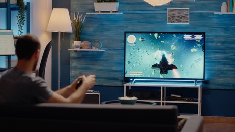 caucasian man playing video games on tv console using joystick while sitting on couch in living room. young person holding controller for entertainment while gaming and enjoying activity