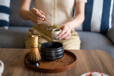 woman making japanese matcha green tea beverage at home using traditional tea ceremony bamboo set while sitting on the sofa at home.