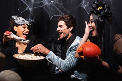 african american woman holding spooky toy hand near interracial friends eating popcorn on halloween party on black