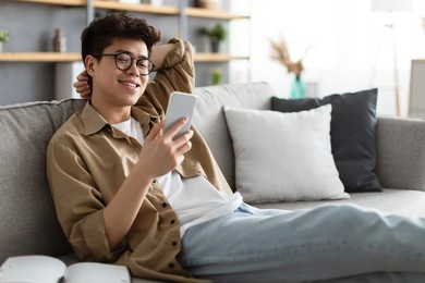 enjoying weekend. handsome asian man in glasses using smartphone while sitting leaning on couch at home, happy guy relaxing in living room, browsing internet or messaging with friends, selective focus