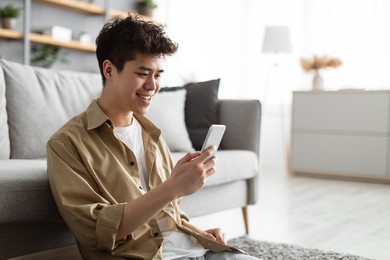 testing new app. handsome asian man using smartphone while relaxing leaning on couch at home, happy guy sitting on floor in living room, browsing internet or messaging with friends, free copy space