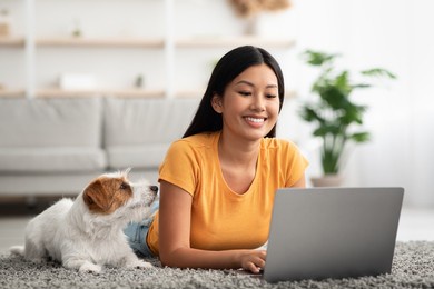 loyal dog looking at its female owner using laptop at home, jack russel terrier puppy laying by happy young asian woman chatting with friends, watching movie online, pet and human concept, copy space