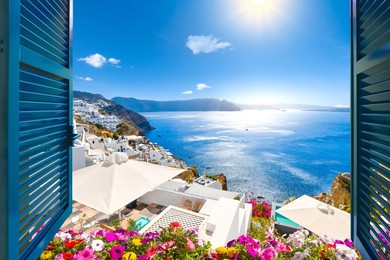 view from an open window with blue shutters of the aegean sea, caldera, coastline and whitewashed town of oia, santorini, greece.
