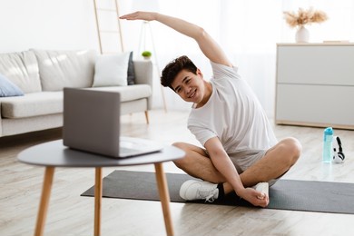 positive athletic asian man stretching arms at home, using laptop, watching sport videos on internet, having fitness class online with coach. healthy lifestyle, sport on self-isolation and quarantine