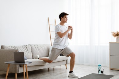 online training. smiling sporty asian guy doing single-leg split squats near couch exercising using laptop computer during workout at home. domestic male fitness and sporty lifestyle. side view