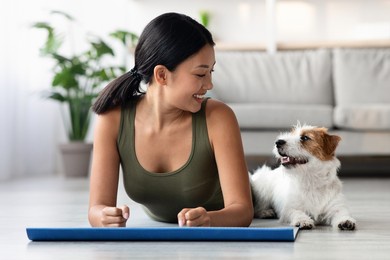 beautiful puppy exercising with its owner athletic asian woman, cheerful young lady in sportswear laying on yoga mat in living room and looking at cute jack russel terrier laying by, copy space