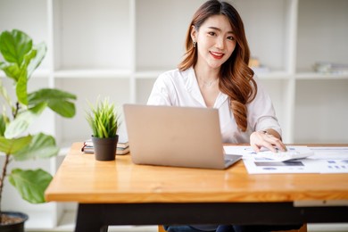 young asian businesswoman beautiful charming smiling and working with a computer laptop in the office. doing financial analysis reports the graph of the company's and growth market research.