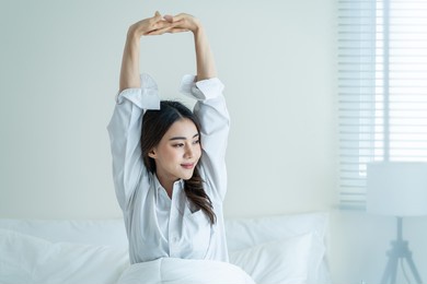 asian beautiful girl in pajamas wake up in the morning with happiness. attractive young woman smiling, feel happy and relax then stretching body after getting up from sleep on bed in bedroom at home.