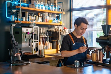 asian man barista using coffee grinder machine grinding roasted coffee beans at cafe. male coffee shop owner brewing black coffee serving to customer. small business restaurant food and drink concept