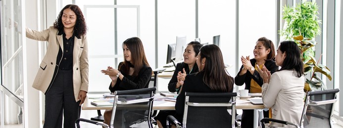 asian middle aged female businesswoman in casual suit standing smiling presenting pointing information on glass board while colleagues sitting at meeting table clapping hands in conference room.