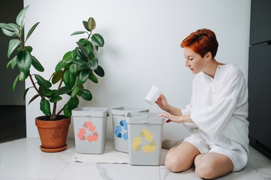 concerned woman sitting on her legs, sorting garbage beween small recycle bins at home. they have differently colored arrows on them. holding plastic cup.