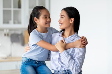 mother daughter love. happy asian lady hugging with her child, sitting in kitchen interior. smiling girl embracing her mom at home, greeting with mother's day, copy space