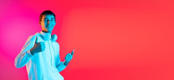 thumbs up, nice sign. portrait of young man, student or boy in casual clothes smiling isolated on magenta studio backgroud in neon. human emotions, facial expression, youth fashion concept. flyer