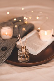 liquid home fragrance in glass bottle with sticks, open paper book, burning scented candles and knit wool textile on tray in bed close up. winter holiday season. cozy atmosphere. aromatherapy. 