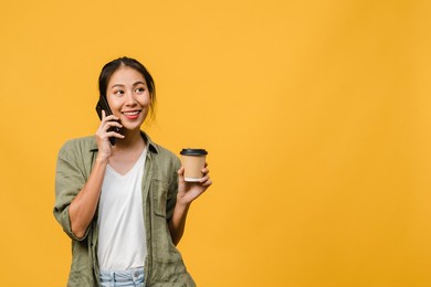 young asia lady talk by phone and hold coffee cup with positive expression, smile broadly, dressed in casual cloth feeling happiness and stand isolated on yellow background. facial expression concept.